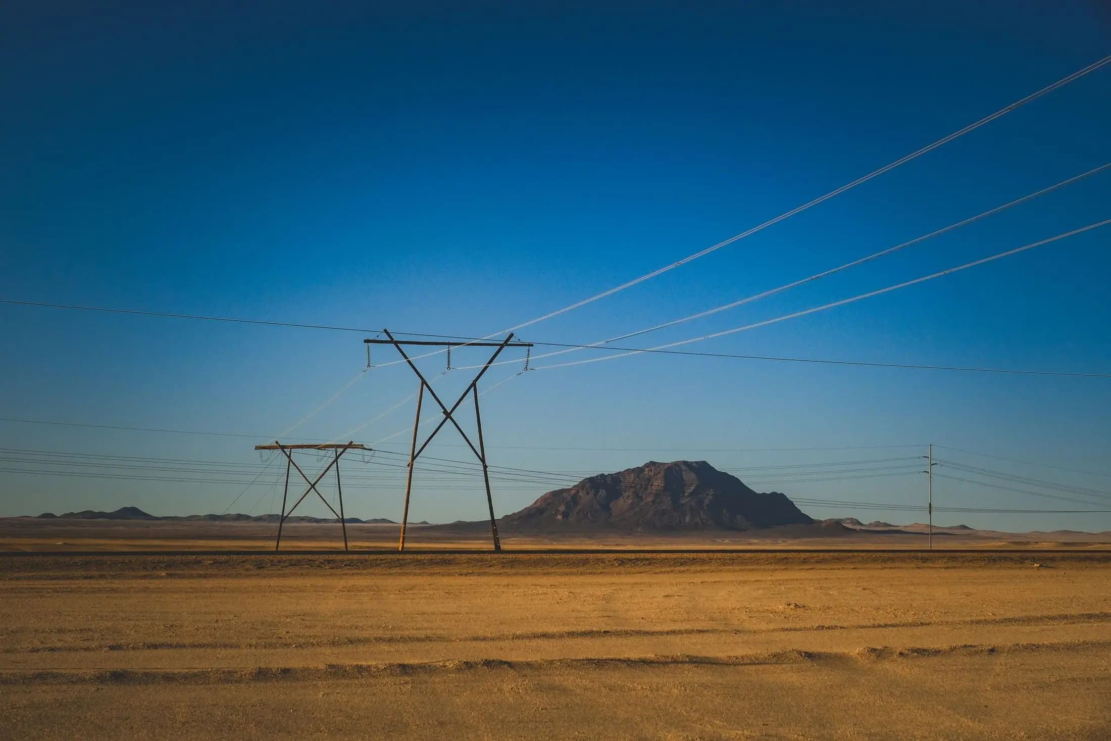 High voltage transmission towers crossing an arid plain toward a lone mountain at blue hour